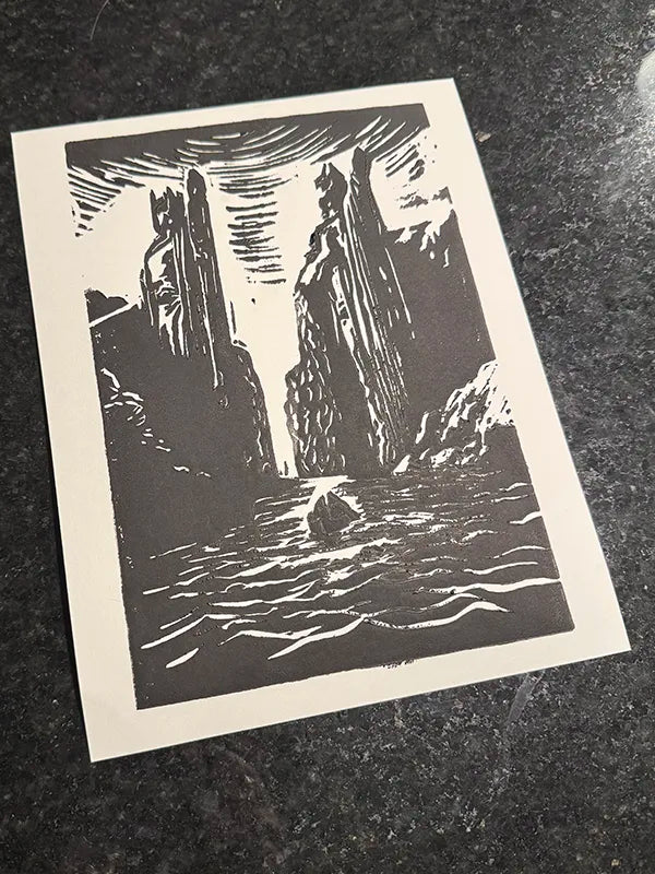 A black and white linocut art print depicting a dramatic canyon scene with a boat on the water, displayed on a dark textured surface.