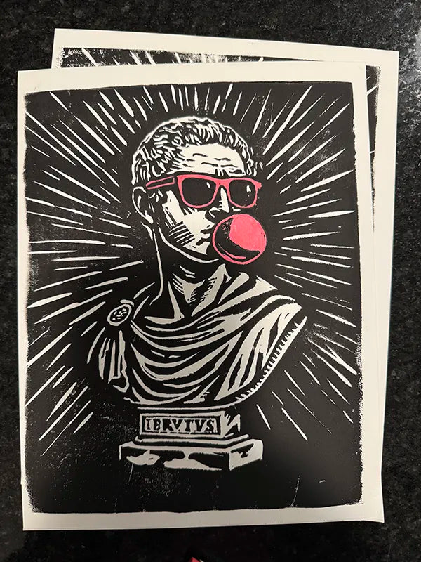 A black and white linocut art print featuring a bust of a Roman-style figure wearing red sunglasses and blowing a pink bubblegum bubble, displayed on a dark textured surface.