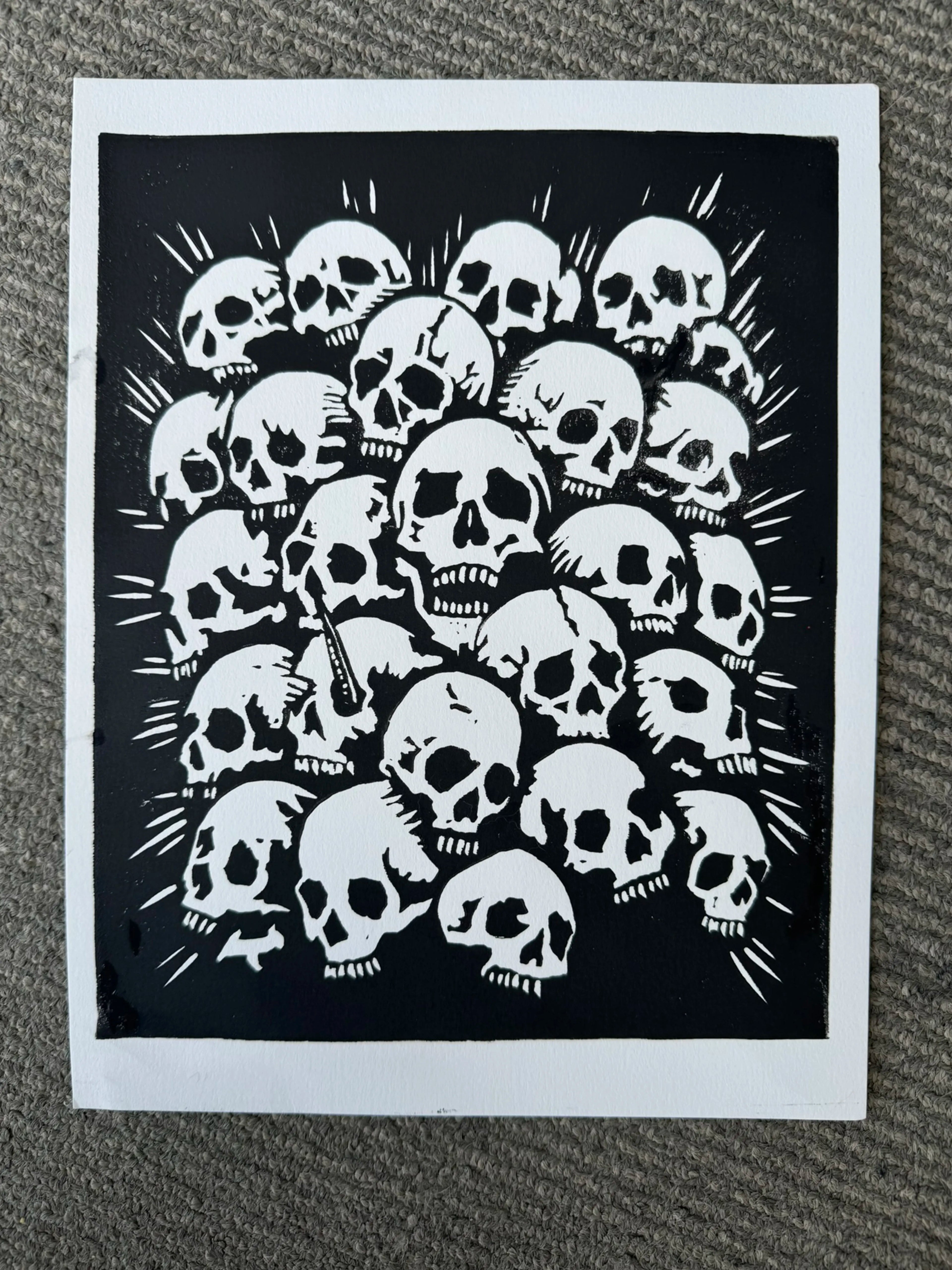 A black and white art print featuring a dense cluster of skulls with radiating lines, presented on a textured gray carpet.