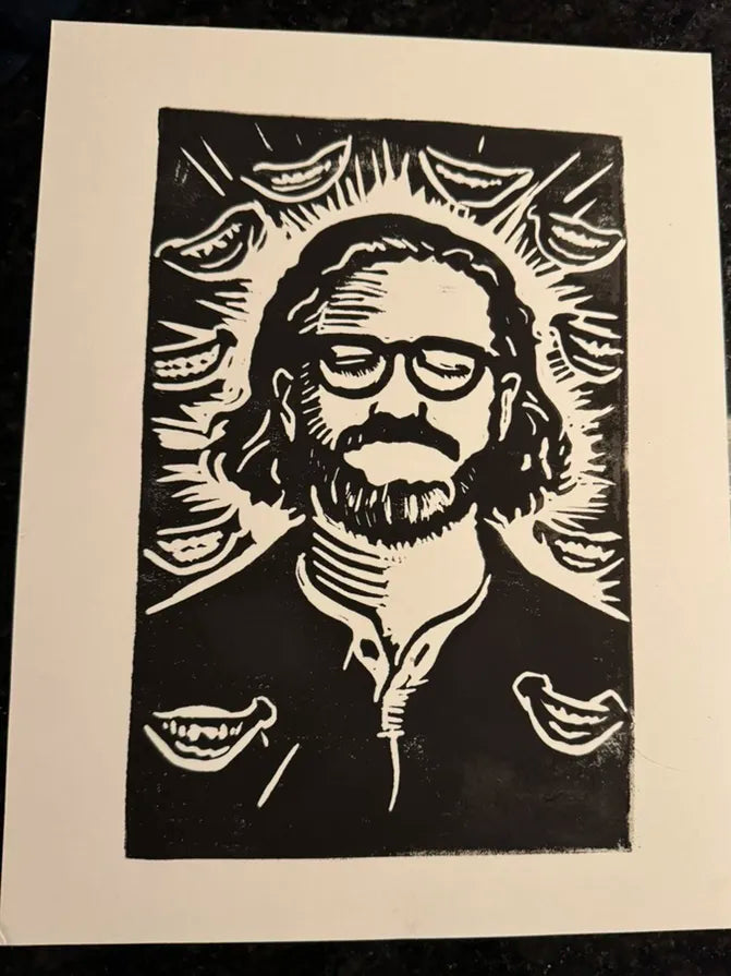 A black and white woodcut-style art print featuring a portrait of a bearded man with glasses, surrounded by multiple smiling faces in a radiating pattern, photographed against a dark textured background.