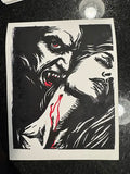 A black and white graphic art print featuring a vampire with red eyes biting down on a woman's neck, with red blood details, displayed on a dark textured surface.