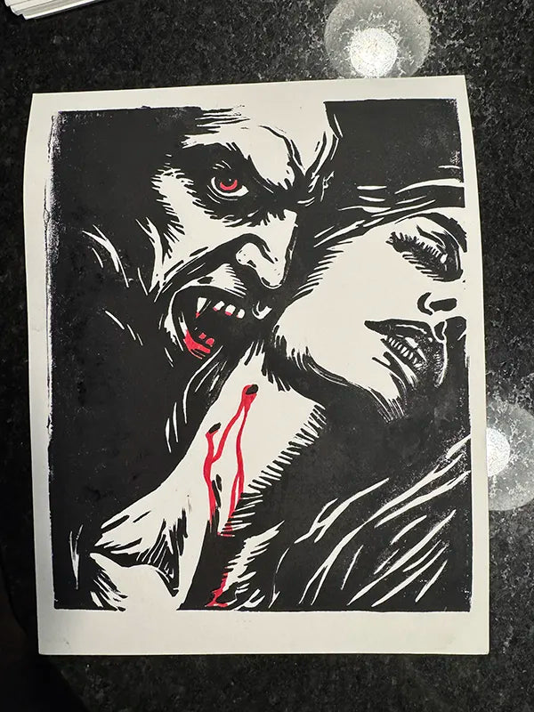 A black and white graphic art print featuring a vampire with red eyes biting down on a woman's neck, with red blood details, displayed on a dark textured surface.